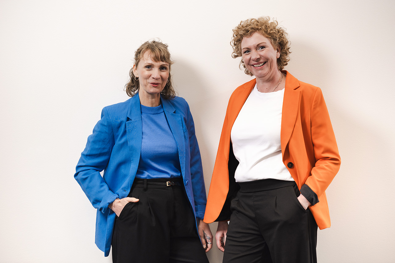im Halbporträt lächelnd an weißer Wand lehnend: Maren Thier und Anja Eglmeyer, Geschäftsführerinnen der designerei